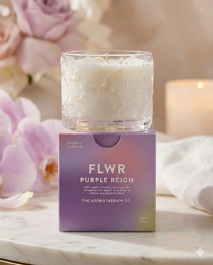The Flwr Purple Reign Candle by The Aromatherapy Company, in a glass jar with a floral design, sits on a purple ombre box surrounded by soft pink flowers and a softly blurred candle in the background.