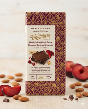 A Whittaker New Zealand Black Doris Plum and Roasted Almonds chocolate bar stands upright on a light surface, surrounded by almonds and plum slices. The packaging is purple and gold with elegant patterns.