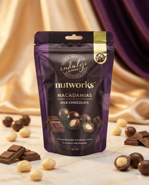 A purple Nutworks Milk Chocolate Macadamias pack stands upright amid whole and halved chocolate pieces and loose macadamias, set against a background of gold satin fabric.