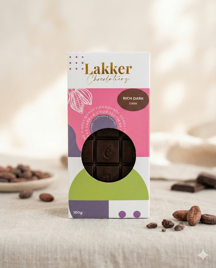 A box of Lakker Chocolatiers Rich Dark Chocolate sits on a light surface, surrounded by cocoa beans and chocolate pieces. The vibrant packaging with geometric shapes showcases the Rich Dark Chocolate bar inside.