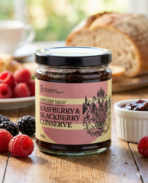 A jar of The Regimental Condiment Company's Raspberry and Blackberry Conserve rests on a wooden table, surrounded by fresh raspberries, blackberries, a slice of bread, and a small dish of jam, with blurred bread and fruit in the background.