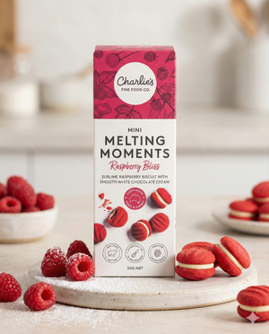 Charlie's Raspberry Bliss Mini Melting Moments Biscuits are shown on a white surface, surrounded by raspberry-filled biscuits, fresh raspberries, and some biscuits dusted with powdered sugar.