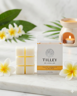 A package of Tilley Tahitian Frangipani Soy Wax Melts rests on a marble surface, surrounded by wax cubes, frangipani flowers, green leaves, and a glowing candle in the background.