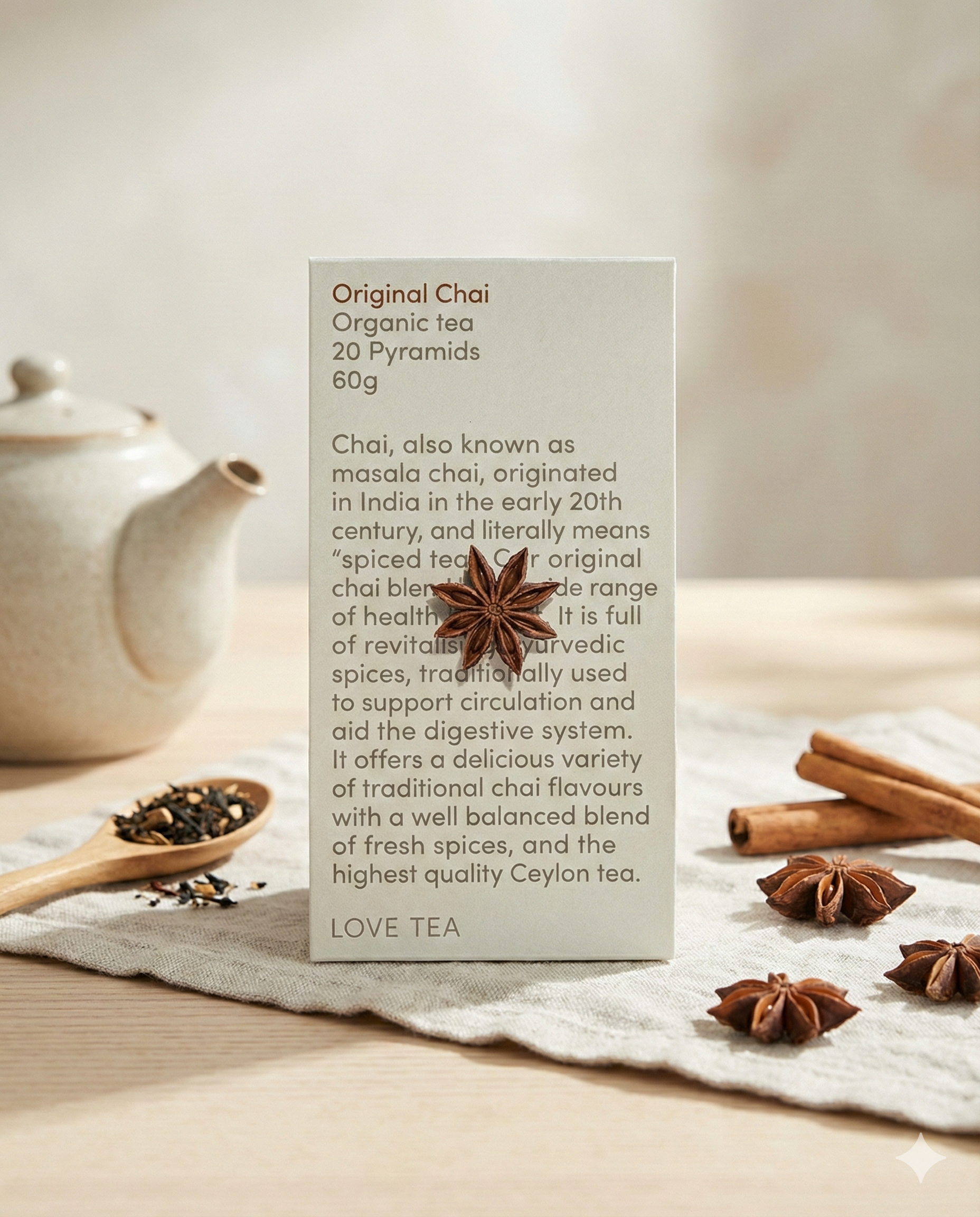 A box of Love Tea Original Chai rests on a wooden surface with star anise, surrounded by a teapot, a bowl of loose tea, and scattered spices like star anise.