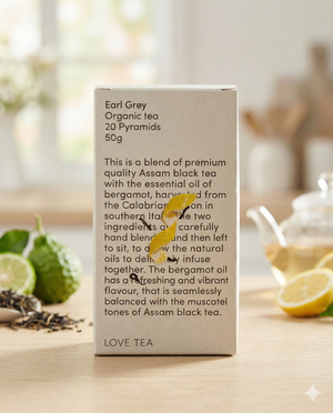 A box of Love Tea's Earl Grey sits on a table with loose tea leaves, a sliced lemon, a teaspoon of tea, and fresh herbs. The box details the Earl Grey blend and its ingredients.