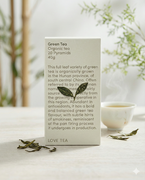 A box of Love Tea Green Tea pyramids sits on a light surface, accompanied by loose green tea leaves and a steaming white teacup. The minimalistic scene highlights the tea’s origin and flavor profile described on the box.