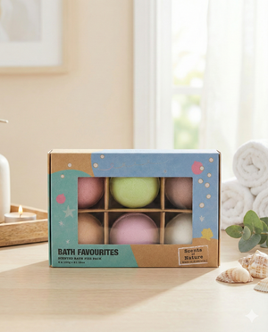 A box of six Tilley LE Bath Favourites Scented Bath Fizz Pack sits on a light wooden surface near a lit candle, stacked white towels, and a small green plant, with sunlight streaming in through the window behind.