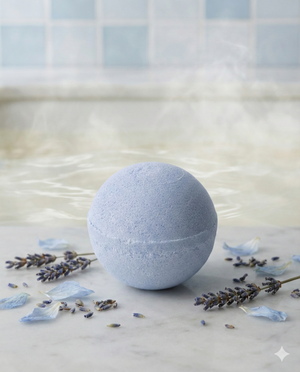 The Tasmanian Lavender Scented Bath Bomb by Tilley rests on marble, surrounded by dried lavender and blue petals, with a steaming bath and tiled wall in the background.