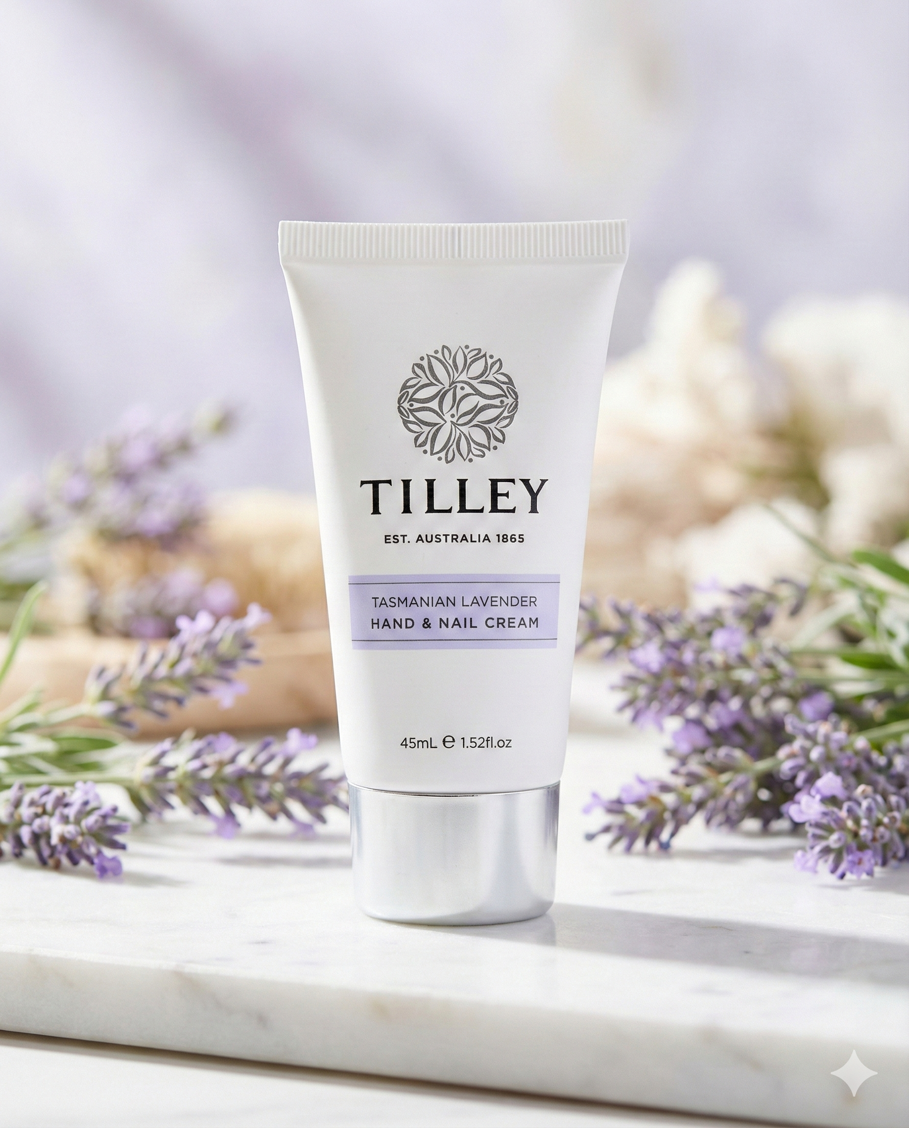 A tube of Tilley Tasmanian Lavender Hand and Nail Cream rests on a marble surface, surrounded by fresh lavender sprigs, with a softly blurred background in neutral and lavender hues.