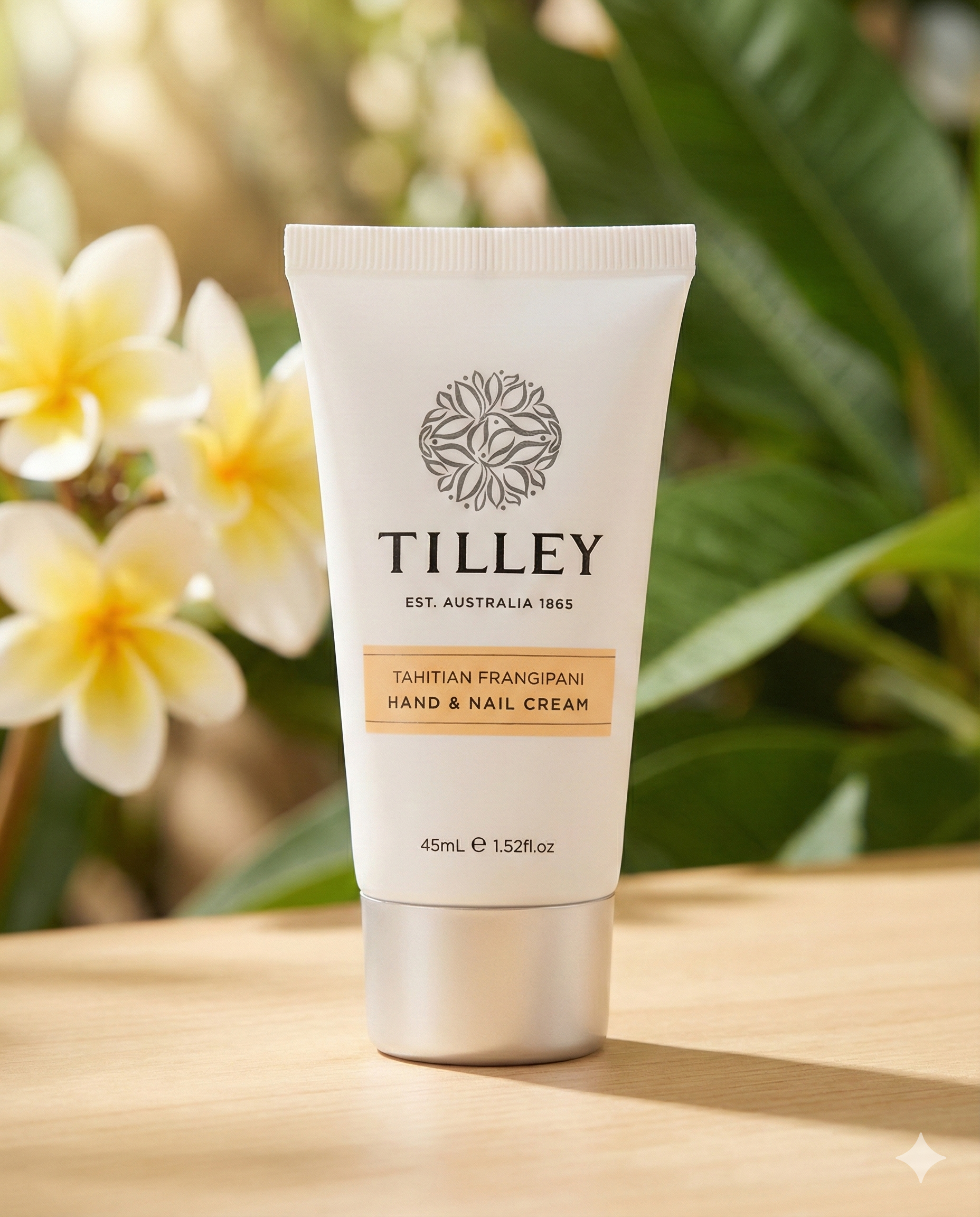 A tube of Tilley Tahitian Frangipani Hand and Nail Cream stands on a wooden surface, with blurred frangipani flowers and green leaves in the background.