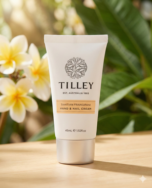 A tube of Tilley Tahitian Frangipani Hand and Nail Cream stands on a wooden surface, with blurred frangipani flowers and green leaves in the background.