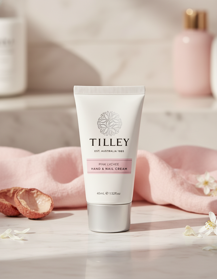 A tube of Tilley Pink Lychee Deluxe Hand and Nail Cream rests on marble with a pink cloth, lychee fruit, white flowers, and softly lit blurred bottles in the background.