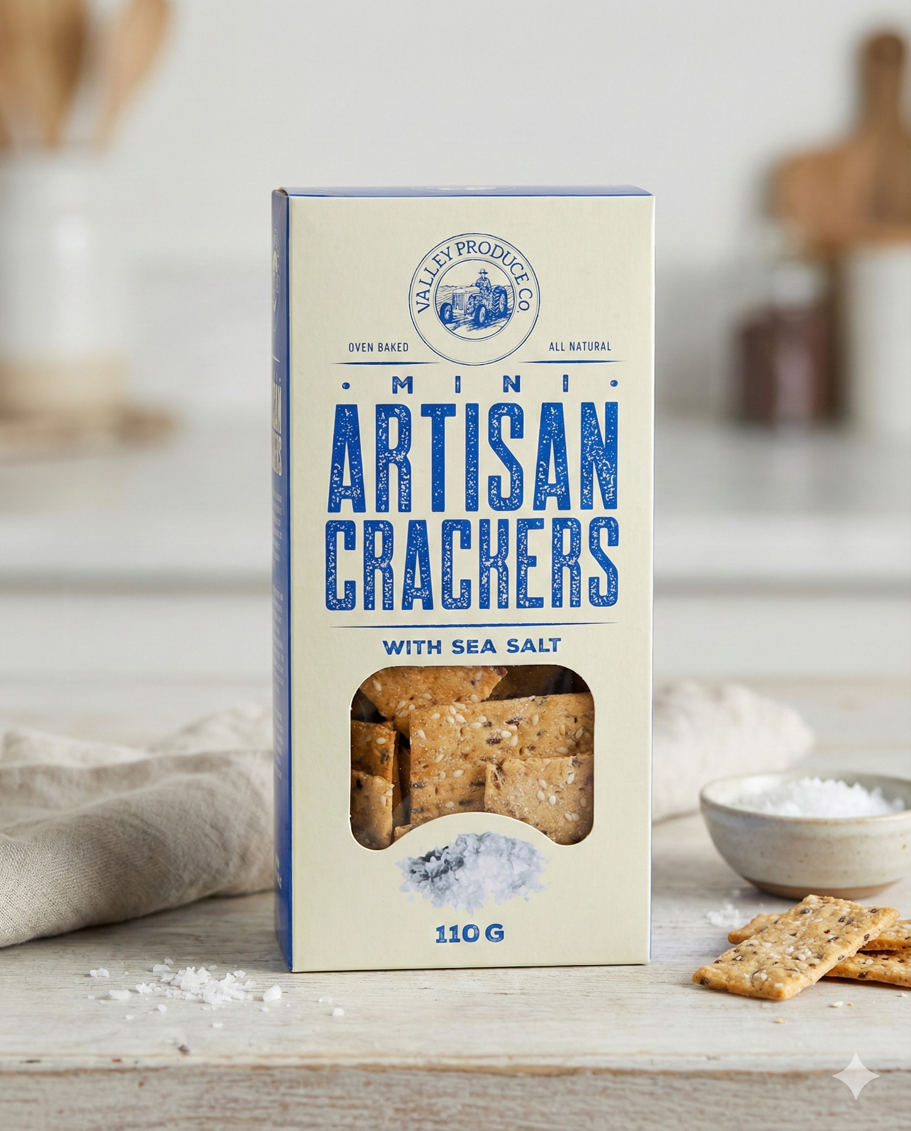 A box of Valley Produce Company Mini Artisan with Sea Salt crackers sits on a light countertop, accompanied by some crackers and a small bowl of sea salt in the foreground, with a softly blurred background.