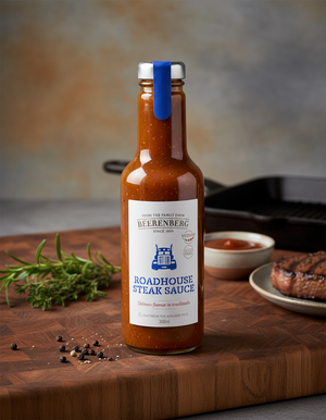 A bottle of Beerenberg Roadhouse Steak Sauce sits on a wooden board, accompanied by rosemary, peppercorns, a small dish of sauce, and a cooked steak in the background.