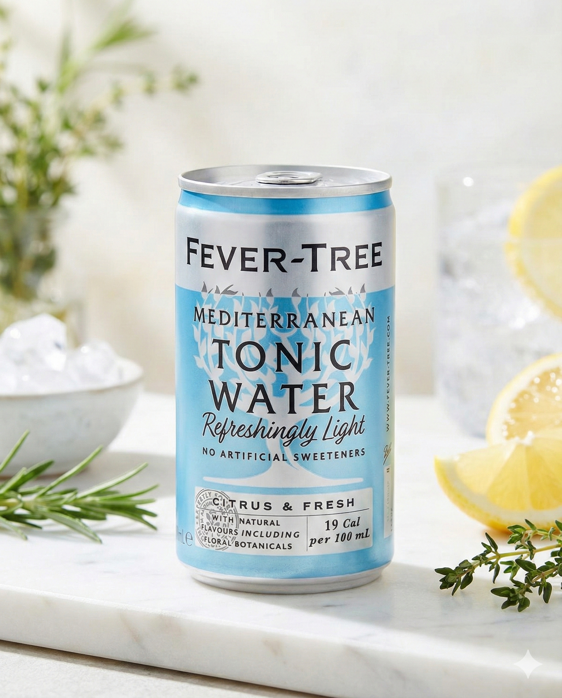 A can of Fever-Tree Mediterranean Tonic Water rests on a marble surface with lemon slices, fresh herbs, and an ice-filled glass. The can features light blue, white, and silver accents.