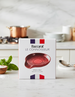 A Baccarat Spoon Rest box sits on a marble counter, with a copper pot and kitchen utensils in the background. The box displays a red spoon rest and features the French flag colors.