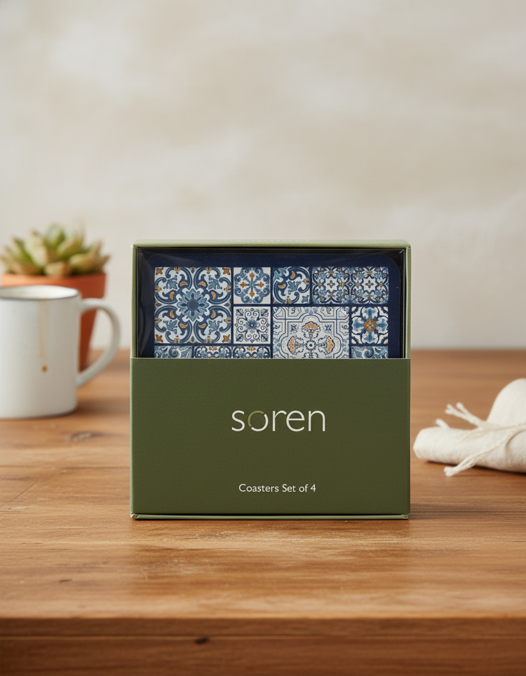 A boxed set of four Soren Coasters featuring blue and white tile patterns sits on a wooden table. The green packaging displays "Soren Coasters Set of 4," with a mug, plant, and napkin in the background.