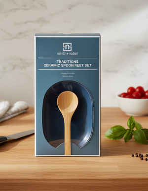 A Smith and Nobel Spoon Rest sits on a wooden countertop with a wooden spoon inside, surrounded by fresh basil, tomatoes, and a kitchen towel.
