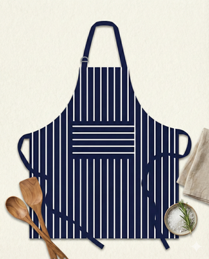 The Smith and Nobel Apron, navy blue with vertical white stripes and a front pocket with horizontal stripes, is displayed on a light surface alongside wooden utensils, a bowl of salt, rosemary, and a folded napkin.