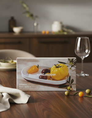 A Salt & Pepper Gibson Platter with yellow, red, and orange heirloom tomatoes sits on a wooden table, accompanied by a glass, napkin, and tomatoes on branches in the background.