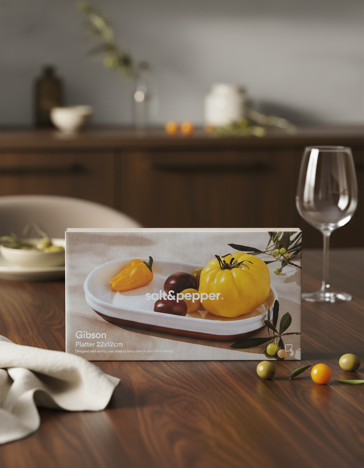 A Salt & Pepper Gibson Platter with yellow, red, and orange heirloom tomatoes sits on a wooden table, accompanied by a glass, napkin, and tomatoes on branches in the background.