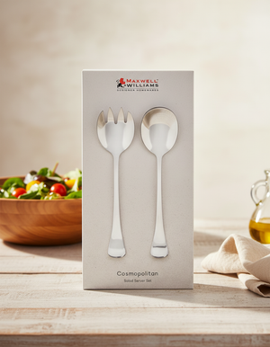 A boxed Maxwell and Williams Cosmopolitan Salad Server Set, with a fork and spoon, is displayed on a table alongside a salad bowl, olive oil, and a folded beige napkin in the background.