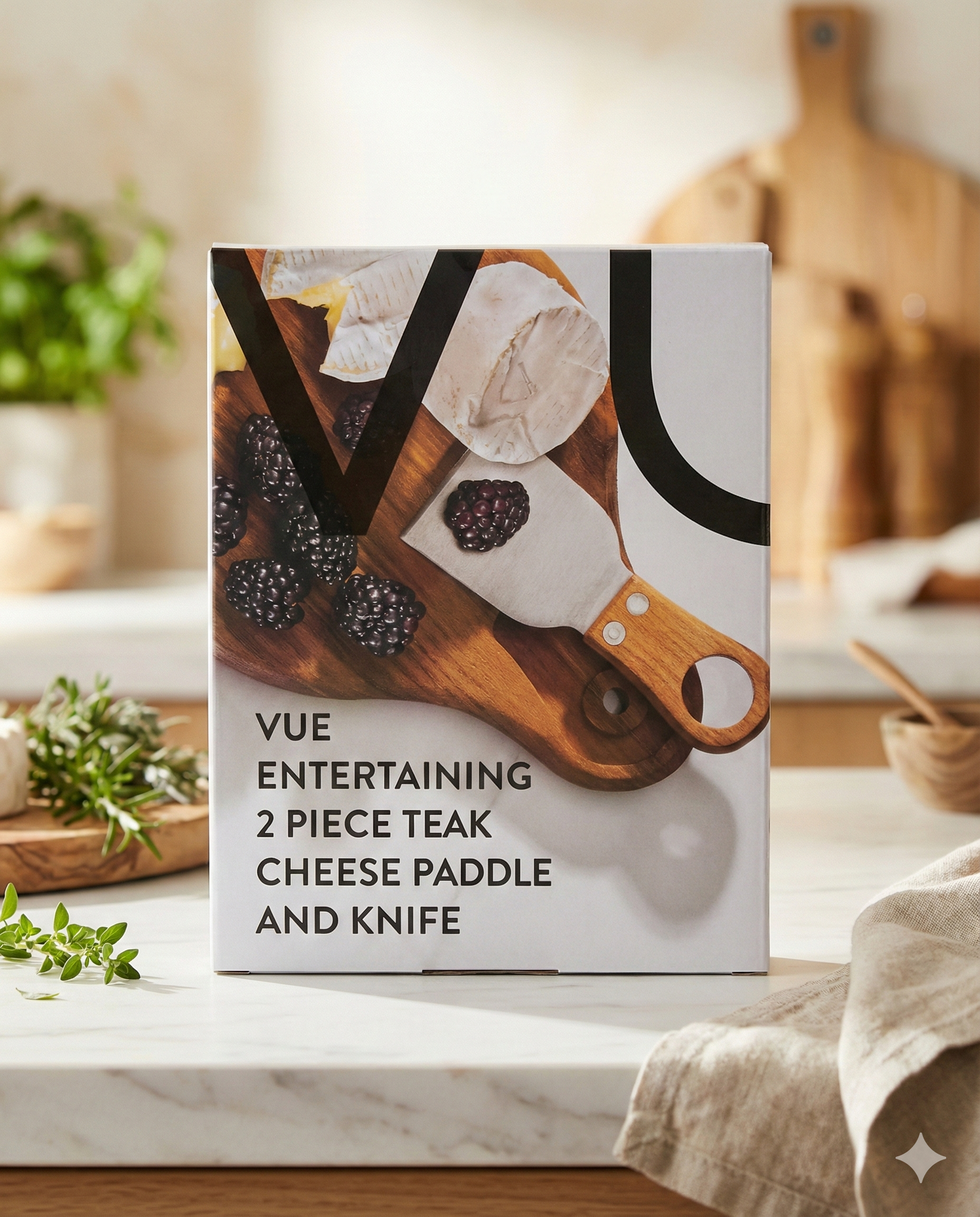 A box of Vue Entertaining Cheese Paddle and Knife sits on a kitchen counter, displaying an image of cheese, blackberries, and the knife on a wooden board, with fresh herbs and kitchen items in the background.