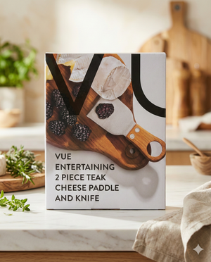 A box of Vue Entertaining Cheese Paddle and Knife sits on a kitchen counter, displaying an image of cheese, blackberries, and the knife on a wooden board, with fresh herbs and kitchen items in the background.