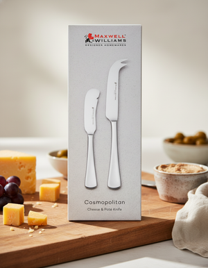 A Maxwell and Williams Cosmopolitan Cheese and Pate Knife is displayed upright on a wooden board, surrounded by cheese cubes, grapes, a bowl of olives, and a small bowl of dip.