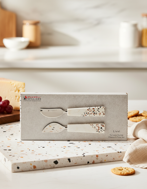 The Maxwell and Williams Livvi Cheese and Pate Set, featuring two cheese knives with terrazzo-patterned handles and a matching cutting board, is displayed in a modern kitchen alongside cheese, grapes, and cookies.