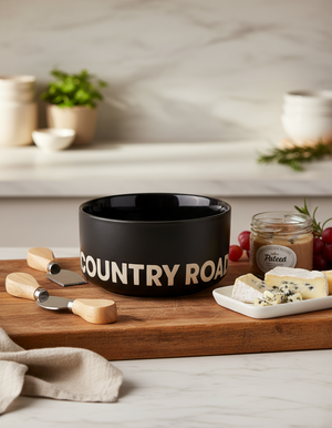 A Country Road Bowl sits on a wooden board with cheese knives, a jar of pâté, grapes, and assorted cheeses in a bright kitchen setting.