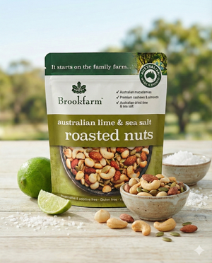 A pouch of Brookfarm Australian Lime and Sea Salt Roasted Nuts is on a table with a bowl of mixed nuts, sea salt, and fresh lime, set against a blurred outdoor background.