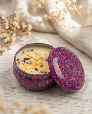 The Gifting Studio Scented Candle with Dried Flowers, featuring a purple mandala decorative tin, is shown partially opened on a light wooden surface, topped with dried flower petals and surrounded by beige fabric and dried flowers.