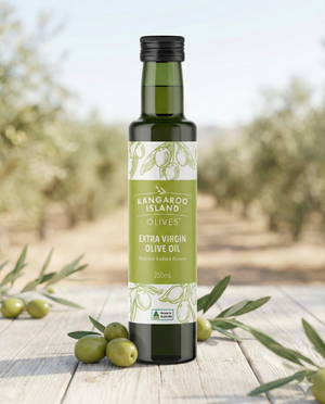 A bottle of Kangaroo Island Extra Virgin Olive Oil is displayed on wood with fresh green olives and olive branches, set before a softly blurred olive grove backdrop.