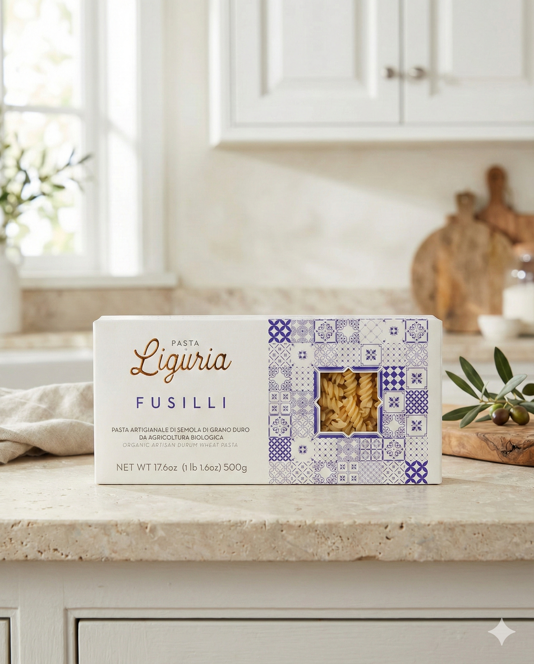 A box of Liguria Fusilli Pasta with white and blue packaging and a clear window sits on a kitchen counter beside olive branches and utensils, with a window in the background.