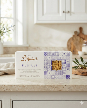 A box of Liguria Fusilli Pasta with white and blue packaging and a clear window sits on a kitchen counter beside olive branches and utensils, with a window in the background.