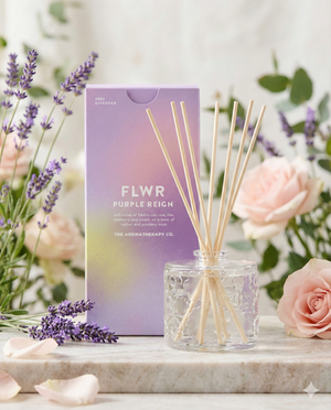 The Aromatherapy Company Flwr Purple Reign Diffuser, featuring light wooden reeds, is placed before a pastel purple box among pink roses and lavender flowers.