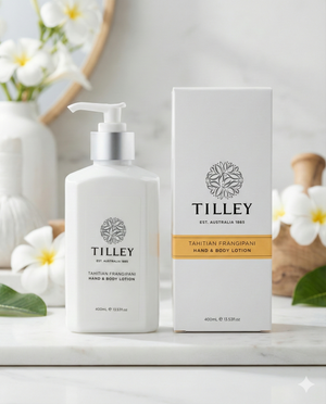 A Tilley Tahitian Frangipani Hand and Body Lotion bottle with a pump and its box are shown on a light surface, surrounded by white and yellow frangipani flowers and greenery.