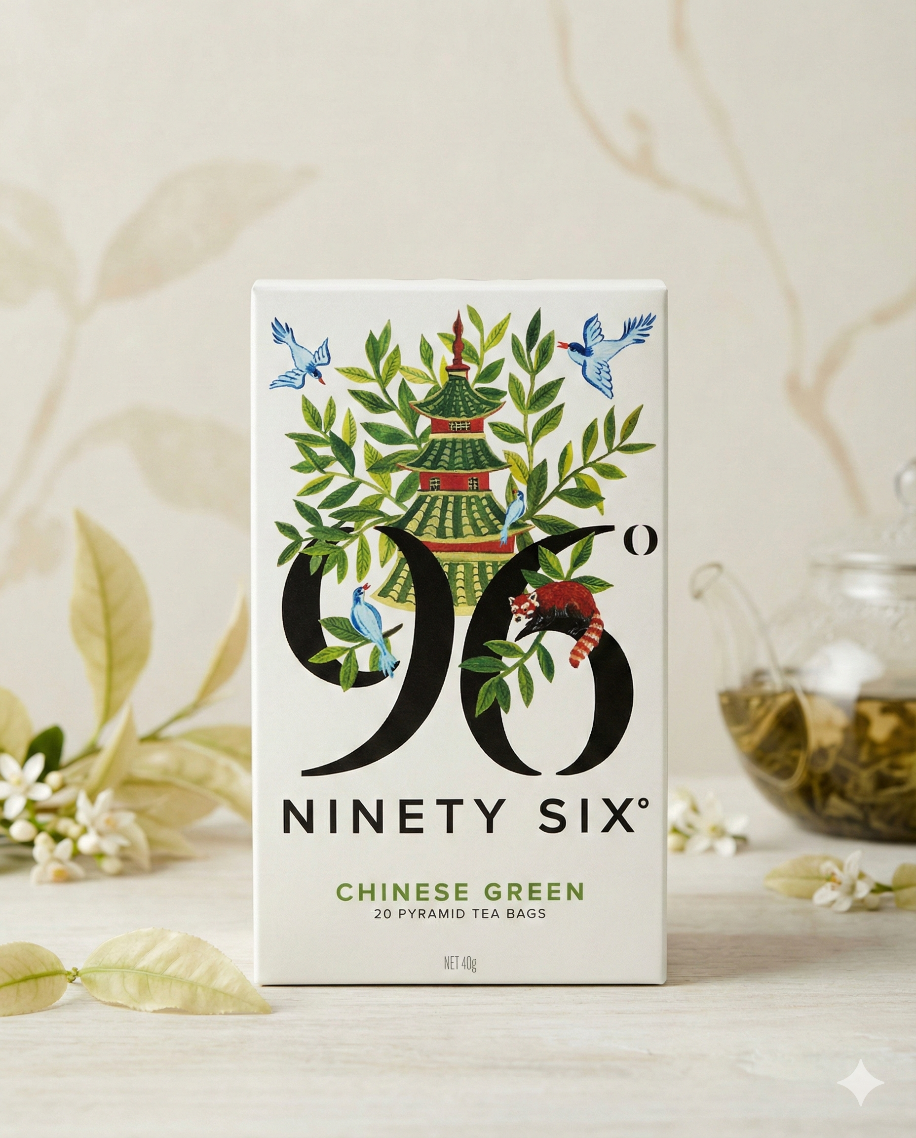 A box of 96 Degrees Chinese Green Tea displays a pagoda, birds, and leaves on the front. In the background are a glass teapot filled with green tea, loose leaves, and light-colored leaves and flowers scattered around.