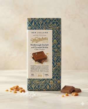 A Whittaker Marlborough Sea Salt and Caramel Brittle bar stands upright on a light surface, surrounded by chocolate pieces, caramel brittle, and sea salt. The blue wrapper features gold geometric patterns.