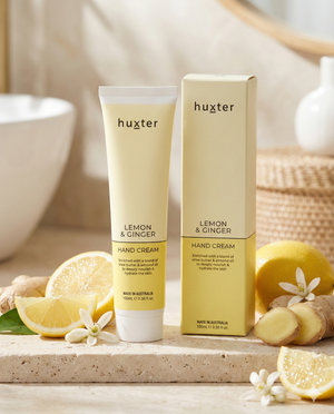 A tube and box of Huxter Lemon and Ginger Hand Cream rest on a beige surface, surrounded by sliced lemons, lemon blossoms, and a woven basket in warm sunlight.