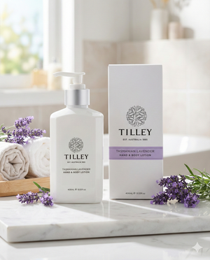A bottle and box of Tilley Tasmanian Lavender Hand and Body Lotion rest on a bathroom counter with sprigs of lavender, two rolled towels, and a softly lit, blurred background.