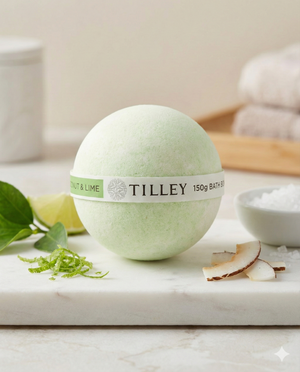 A Tilley Coconut and Lime Scented Bath Bomb rests on marble with lime zest, coconut pieces, a lime wedge, bath salts, and green leaves. Towels and a jar are softly blurred in the background.