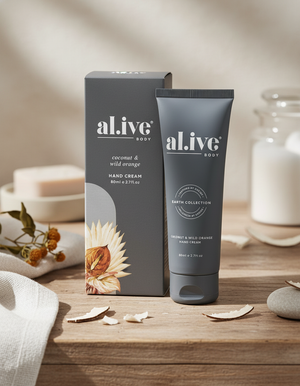 A tube and box of Alive Body Coconut and Wild Orange hand cream are shown on a wooden surface with a towel, dried flowers, a candle, and minimalist decor in soft lighting.