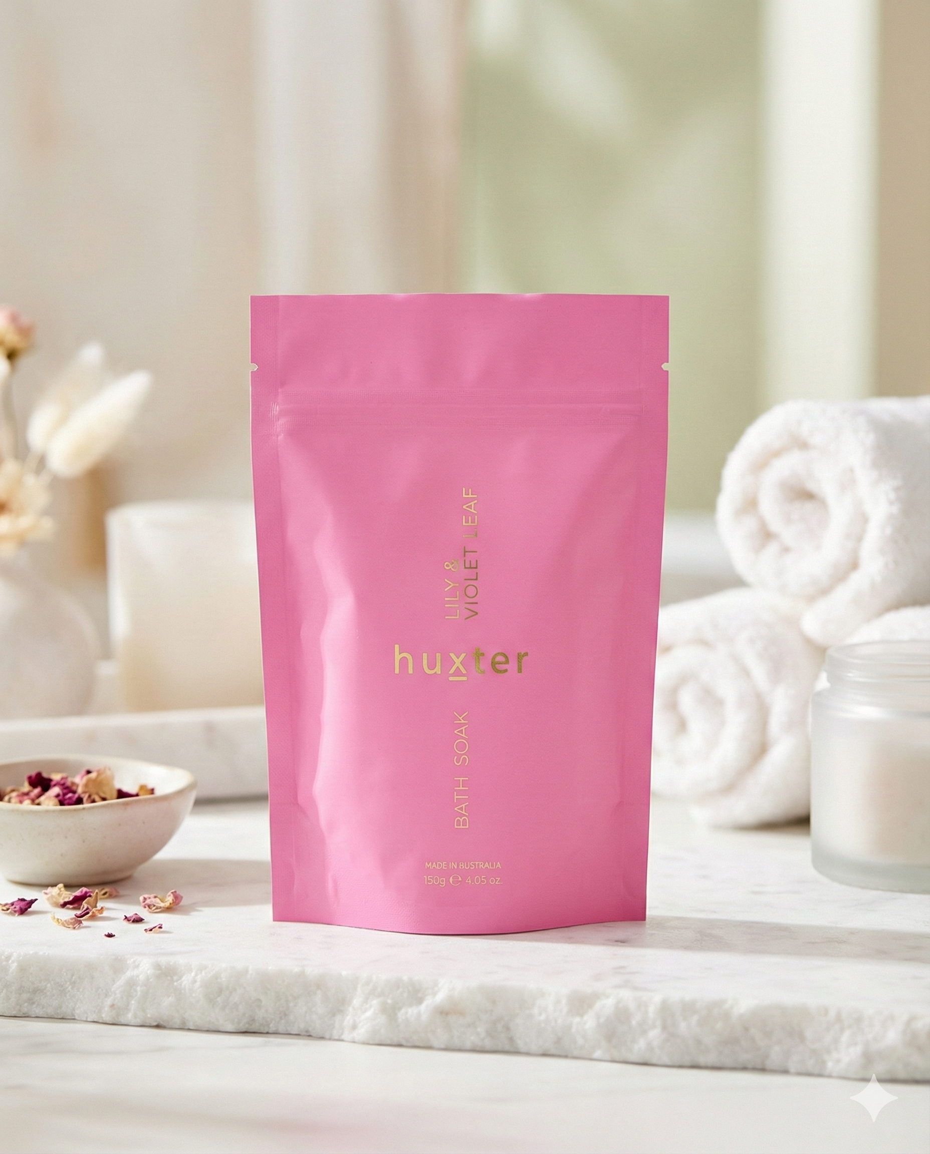 A Huxter Bath Soak Lily and Violet Leaf pouch in pink stands on a white surface, accompanied by rolled white towels, a small bowl of dried flowers, and a blurred bathroom background.