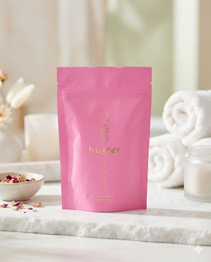 A Huxter Bath Soak Lily and Violet Leaf pouch in pink stands on a white surface, accompanied by rolled white towels, a small bowl of dried flowers, and a blurred bathroom background.