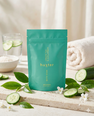 A teal Huxter Bath Soak Green Tea and Cucumber pouch stands on a light surface, surrounded by cucumber slices, green leaves, white flowers, a rolled towel, and a small bowl of bath salts.