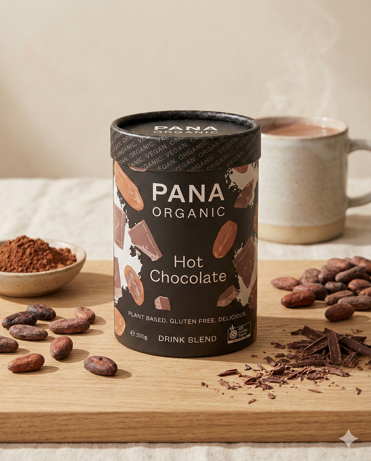 A canister of Pana Organic Hot Chocolate 52% Cacao is displayed on a wooden board with cocoa beans, chocolate pieces, cocoa powder, and a steaming mug of hot chocolate in the background.