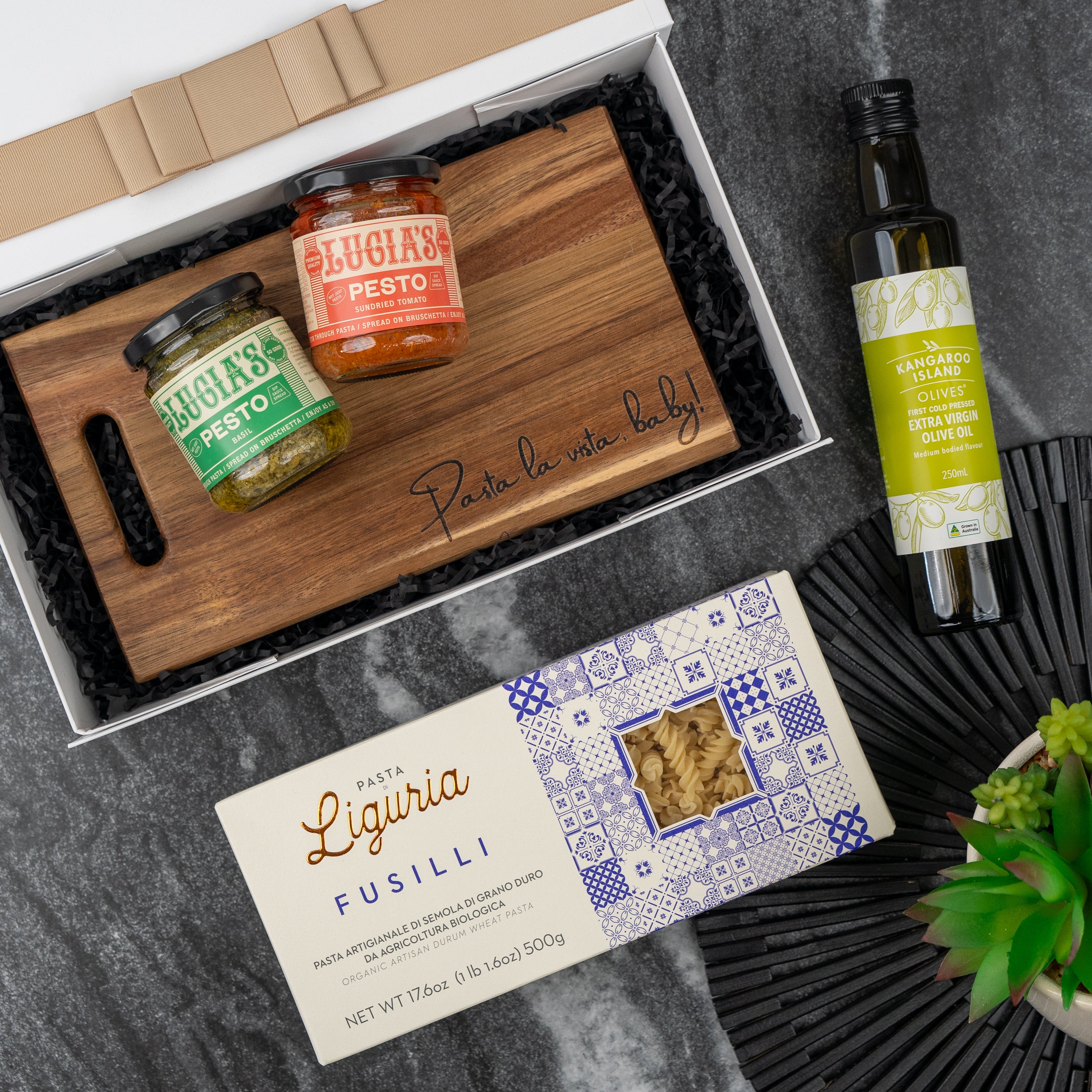 The Hamper Boutique Co Italian Kitchen Hamper includes two jars of pesto, a wooden cutting board, extra virgin olive oil, and Liguria fusilli pasta in a gift box—perfectly displayed beside a green plant on a dark surface.