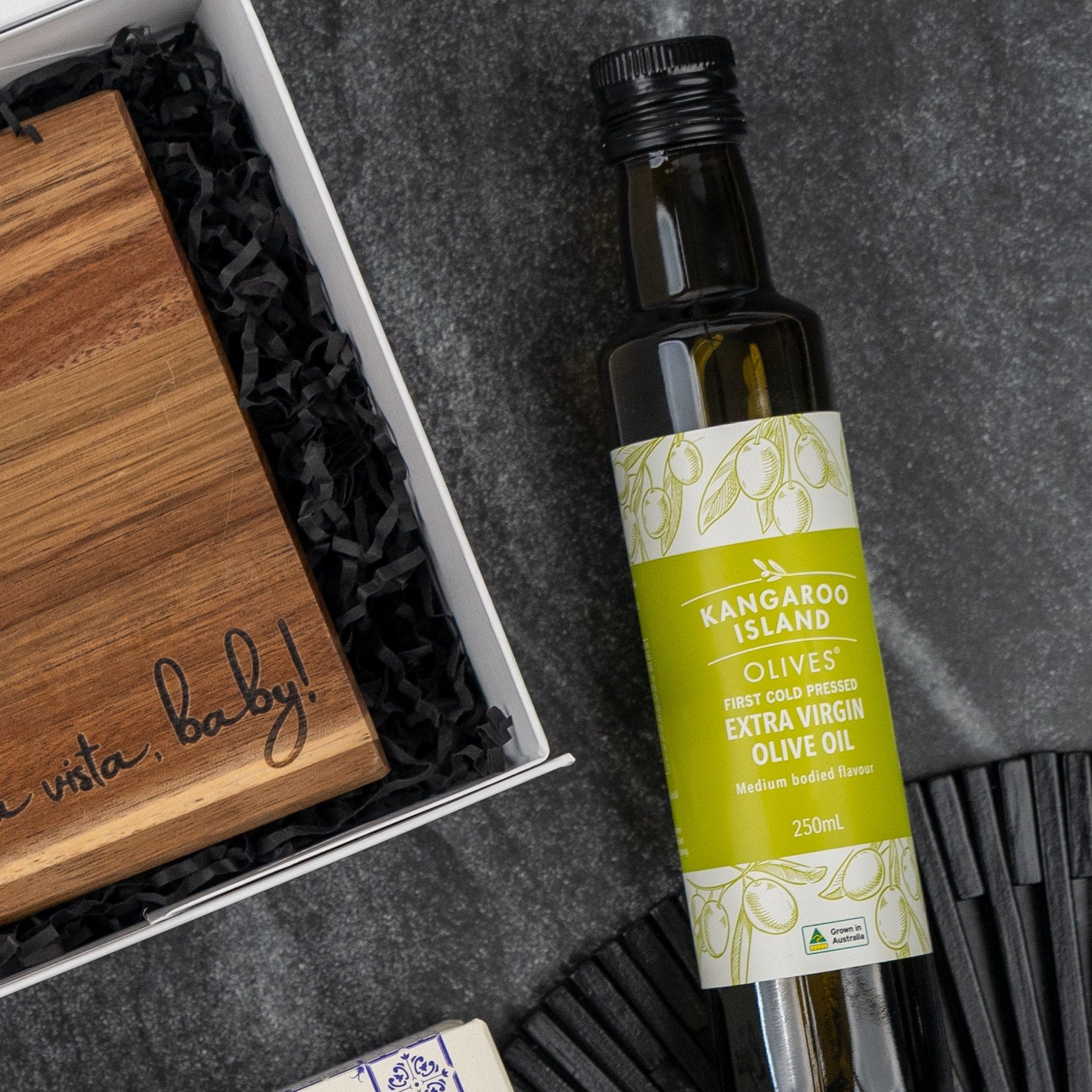 An Italian Kitchen Hamper by The Hamper Boutique Co sits on a textured gray surface with black shredded paper, accompanied by a wooden box inscribed “Hasta la vista, baby!” and a partially visible black item.